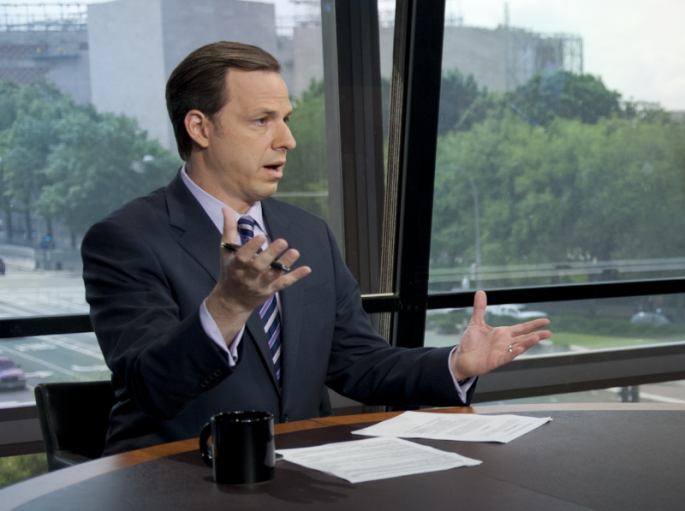 Photograph of Jake Tapper in a dark suit sitting at a news desk with a view of trees and sky scrappers outside a picture window.